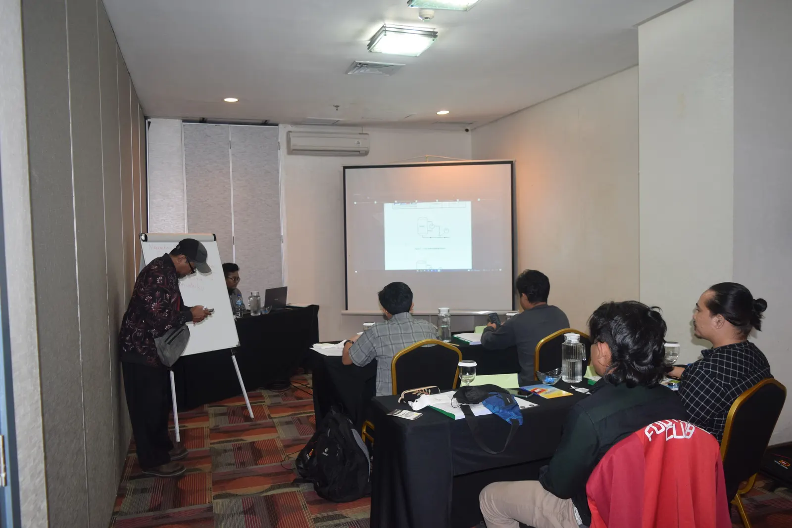 Instruktur menjelaskan diagram teknis di flipchart kepada peserta pelatihan di ruang meeting hotel