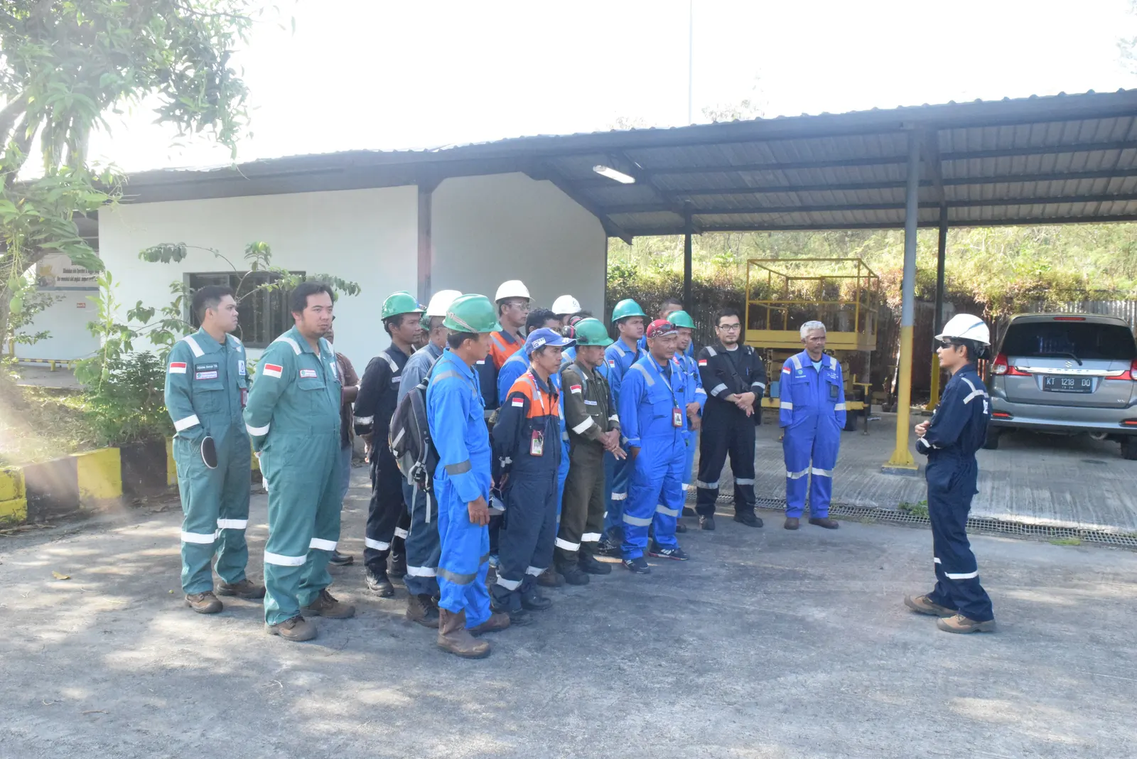 Briefing keselamatan kerja di area luar fasilitas industri oleh instruktur kepada pekerja berseragam coverall dan helm