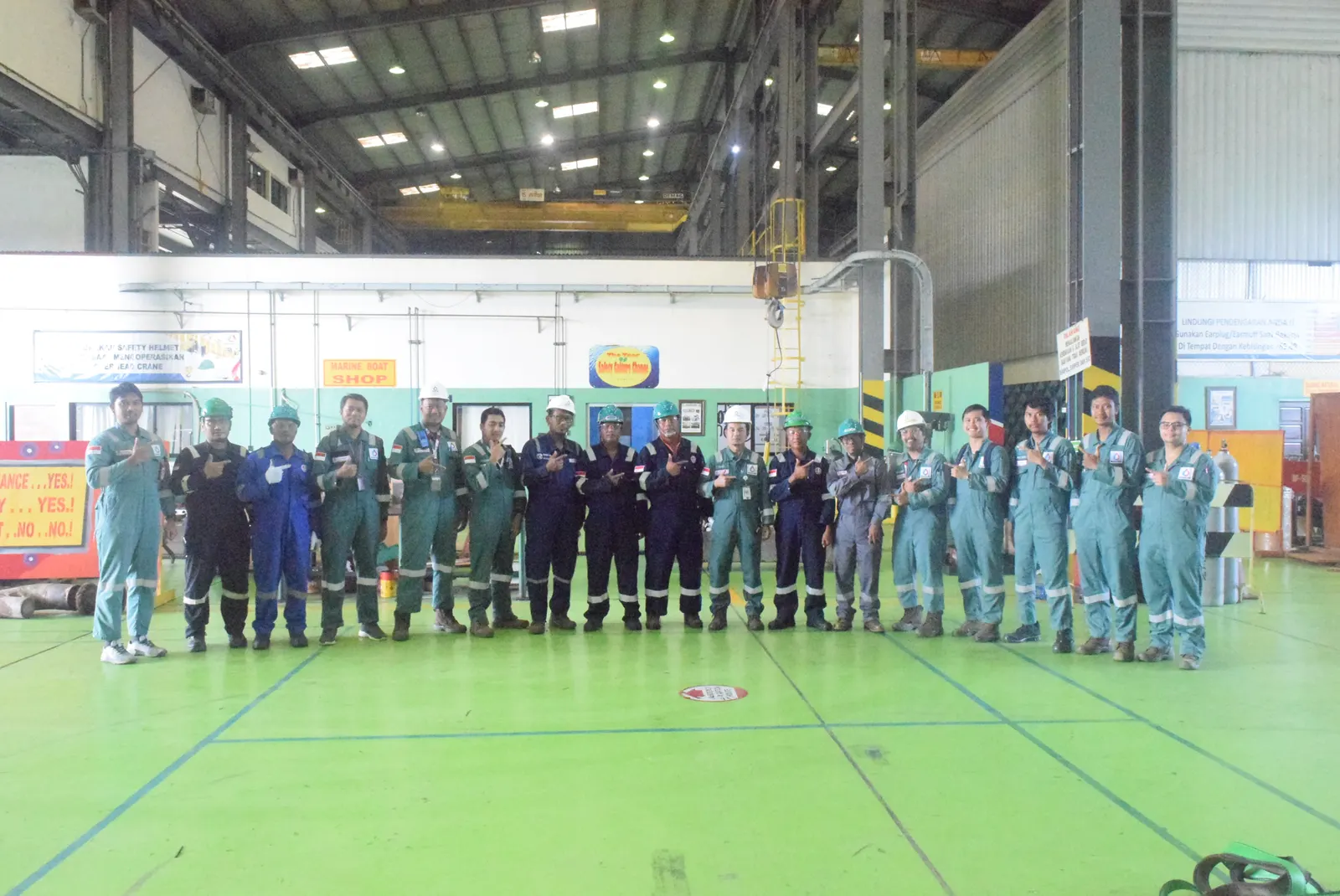 Peserta pelatihan K3 berseragam coverall berfoto bersama di dalam workshop industri dengan overhead crane