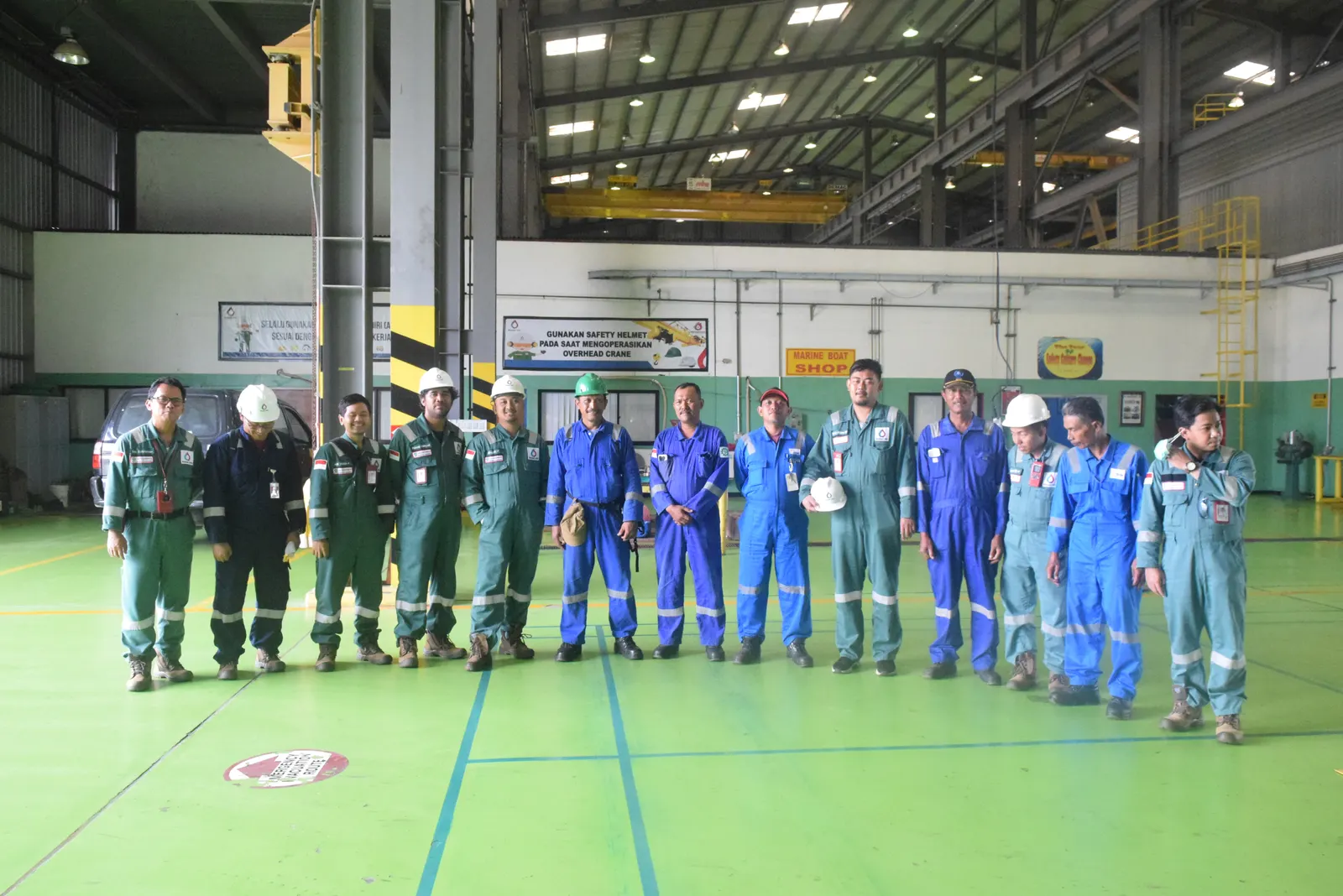 Peserta pelatihan K3 berseragam coverall berfoto di dalam workshop industri dengan spanduk keselamatan