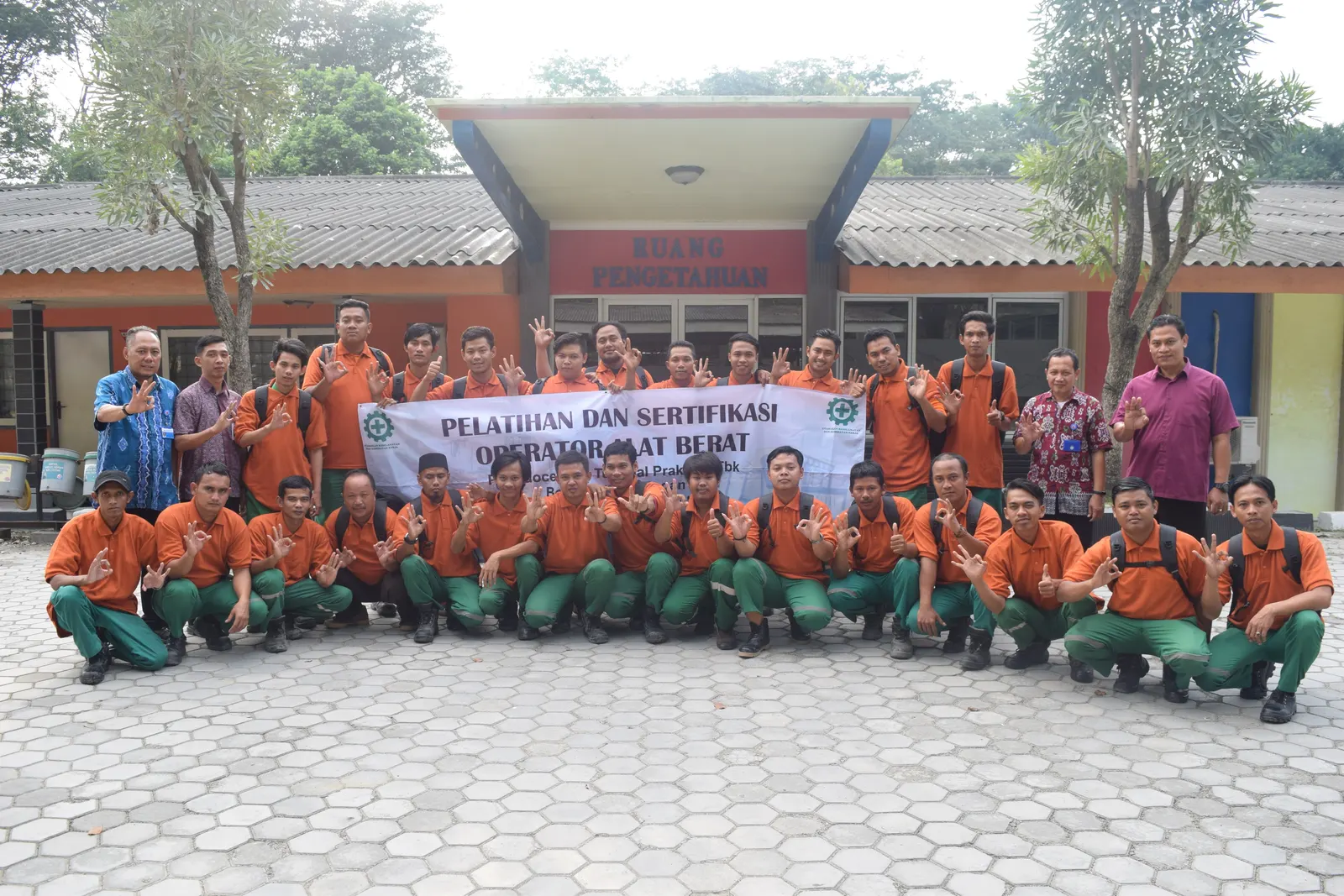 Foto bersama peserta pelatihan dan sertifikasi operator alat berat berseragam oranye-hijau di depan gedung Ruang Pengetahuan