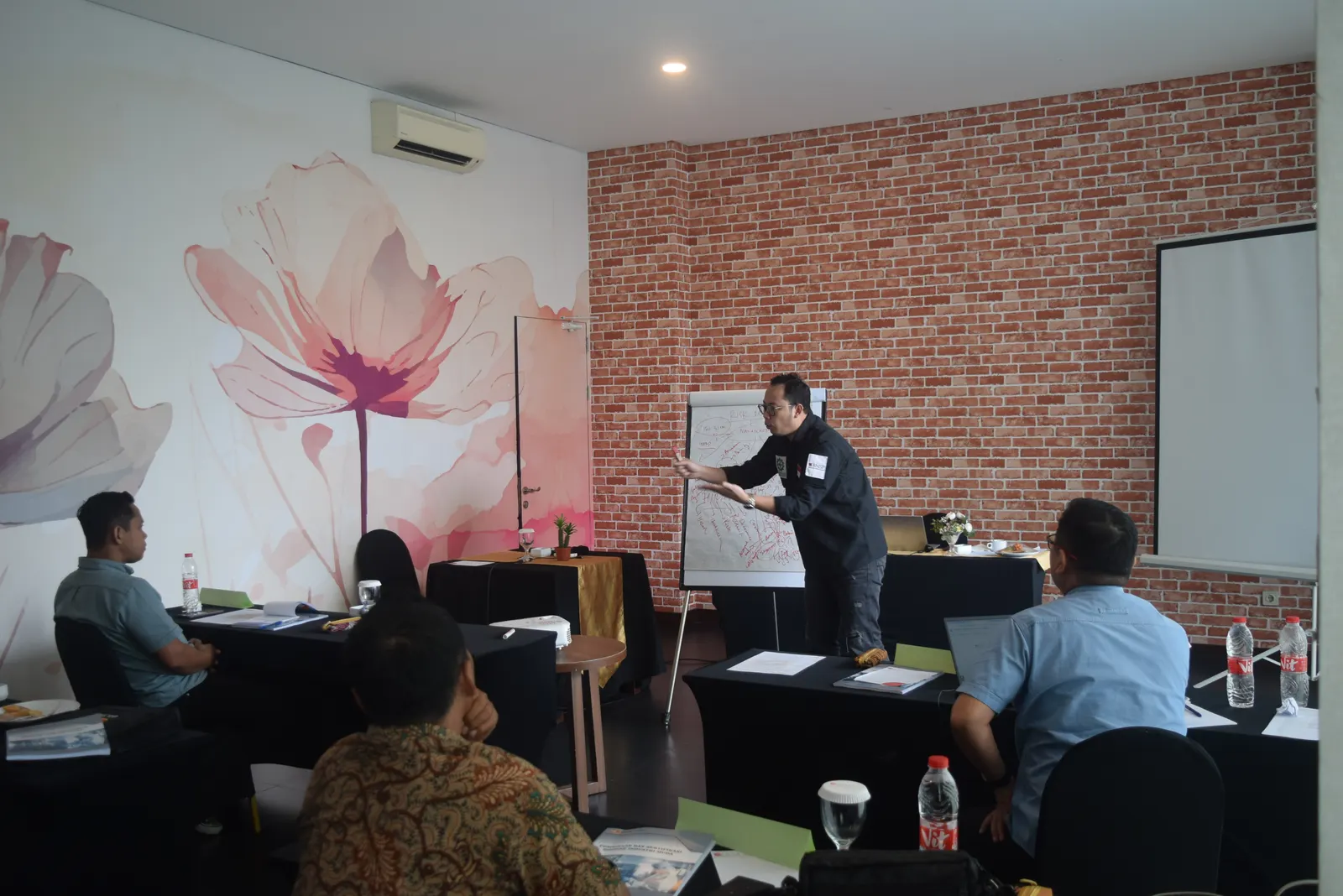 Instruktur menjelaskan materi K3 menggunakan flipchart di ruang pelatihan hotel dengan dinding bata