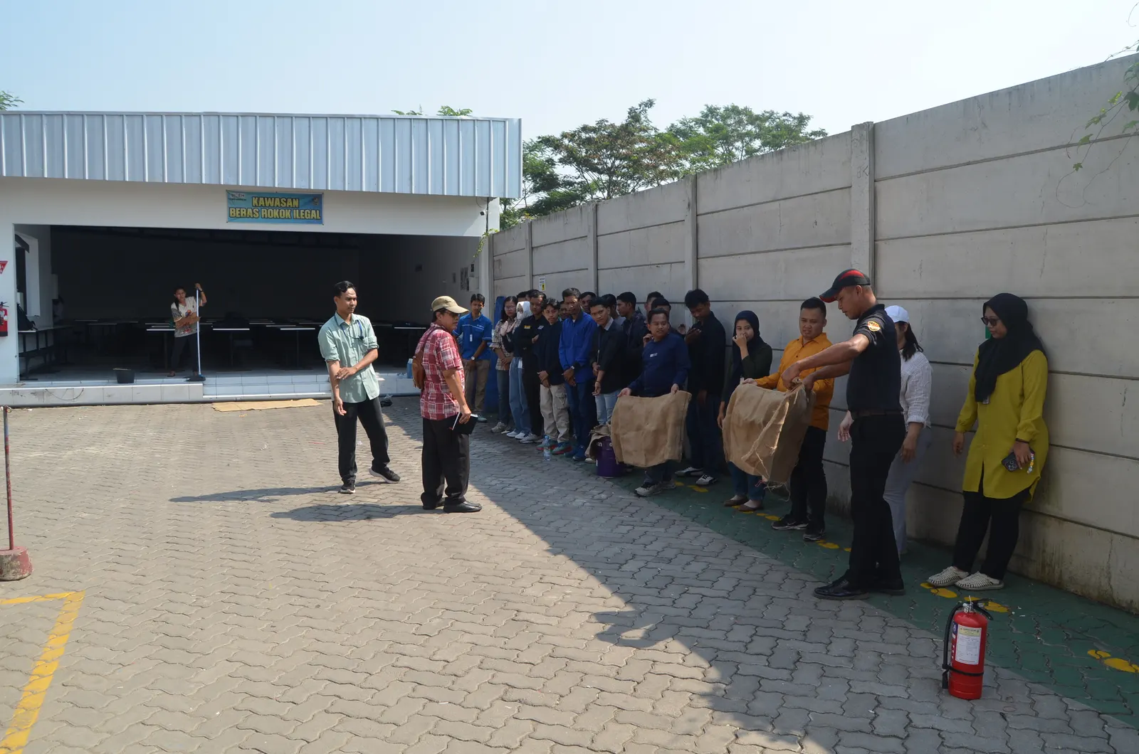 Praktik pemadaman kebakaran dengan APAR di area luar pabrik dengan spanduk Kawasan Bebas Rokok Ilegal