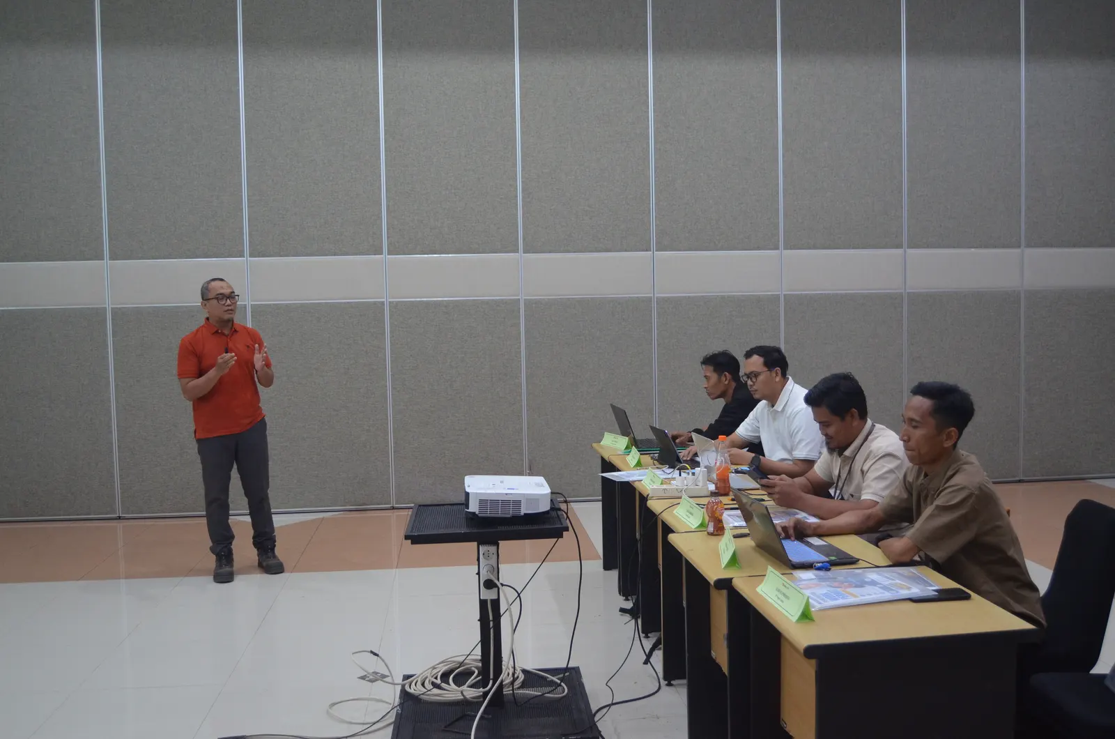 Instruktur berkaos oranye menyampaikan materi kepada peserta pelatihan yang duduk di meja kelas dengan name tag