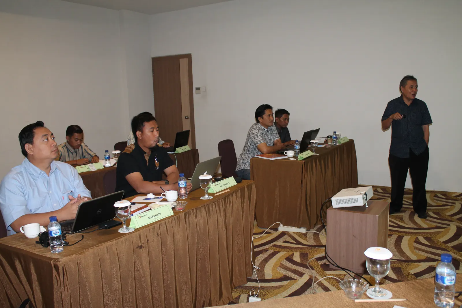 Instruktur menyampaikan materi pelatihan kepada peserta yang duduk di meja kelas hotel dengan name tag dan proyektor