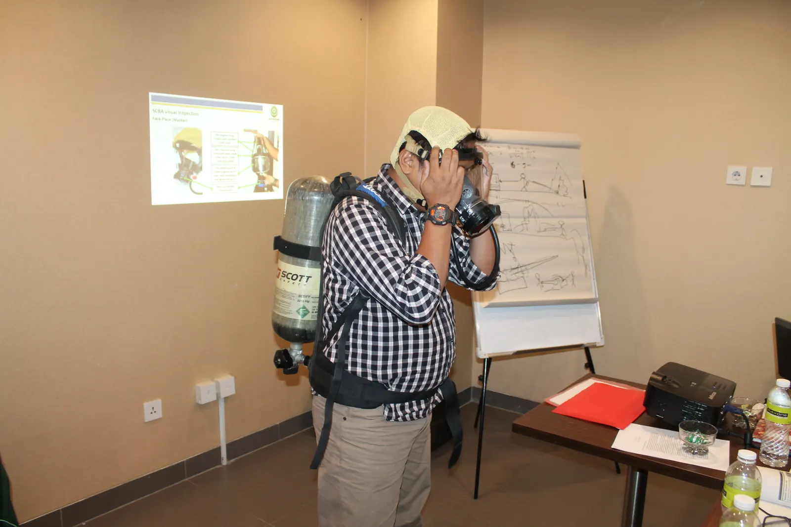 Demonstrasi pemasangan masker SCBA oleh instruktur dengan slide inspeksi visual di latar belakang