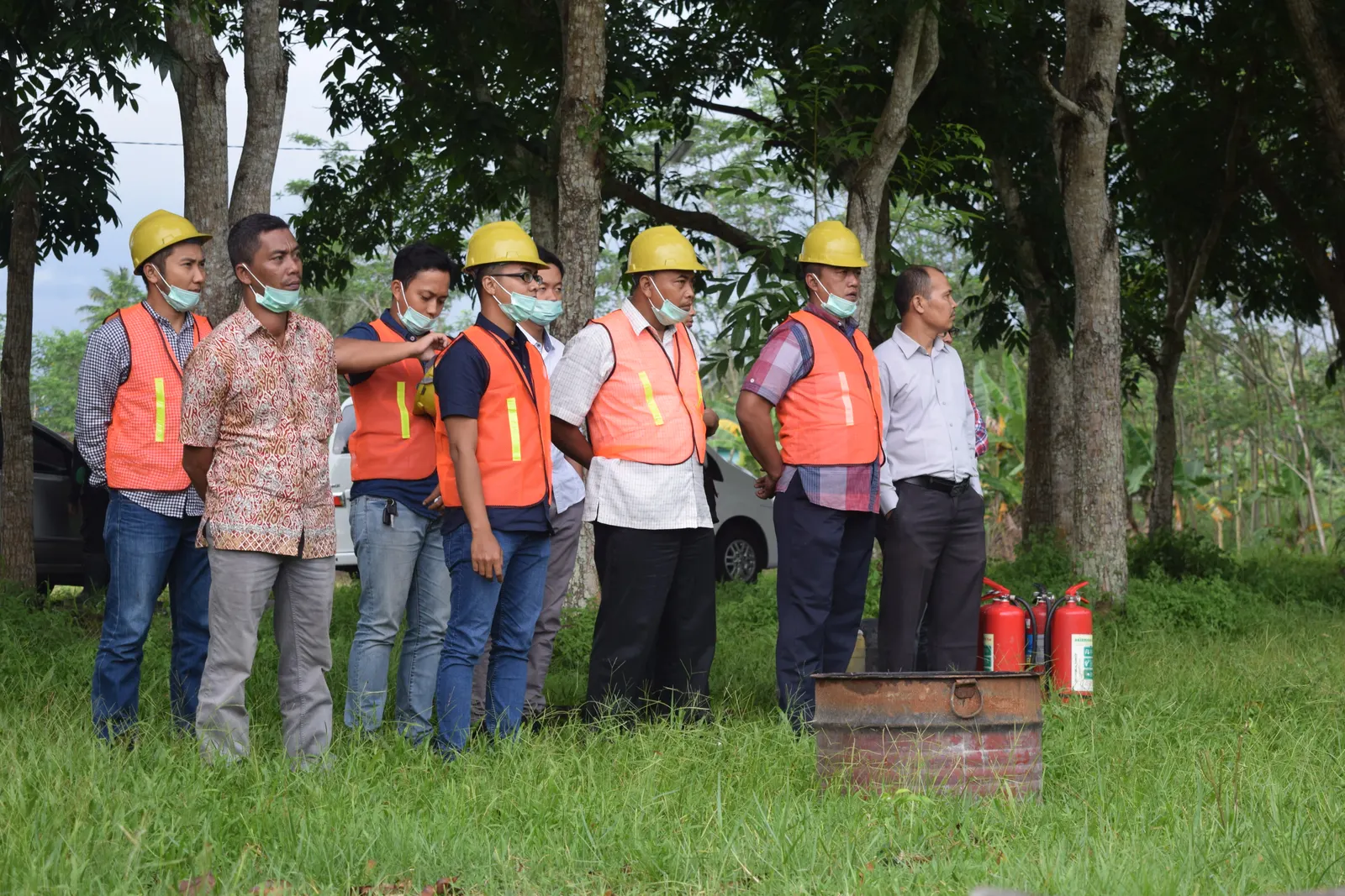Peserta pelatihan K3 berrompi oranye dan berhelm kuning berdiri di dekat alat pemadam kebakaran di area praktik lapangan