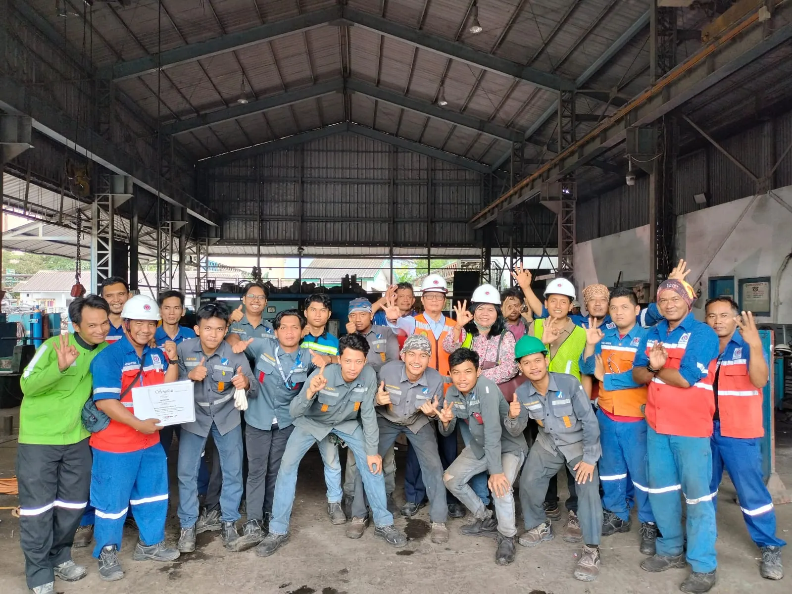 Foto bersama pekerja pabrik dengan sertifikat K3 di dalam workshop industri dengan peralatan berat