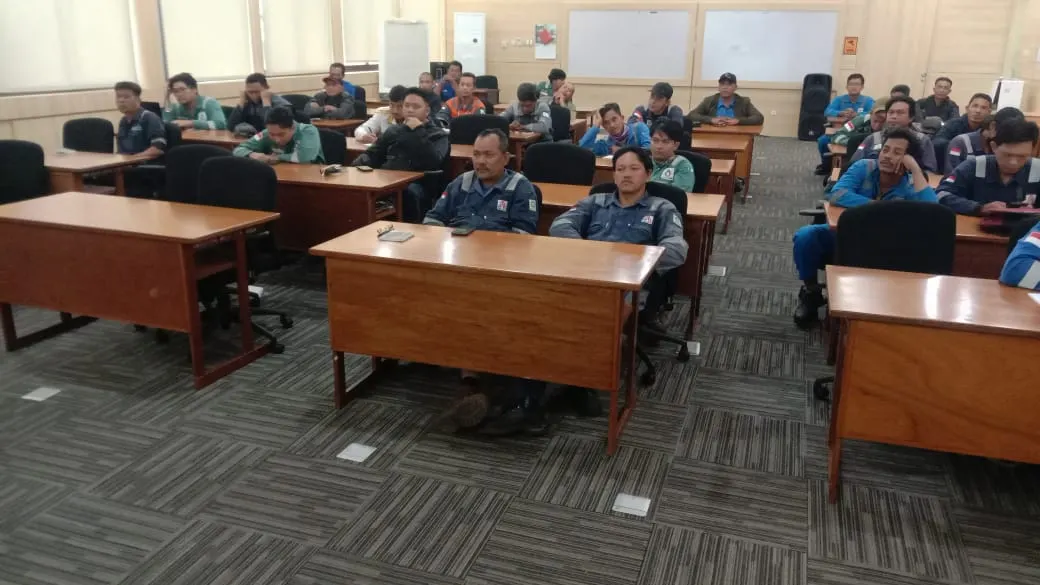 Peserta pelatihan K3 berseragam coverall hijau dan biru duduk di ruang kelas perusahaan